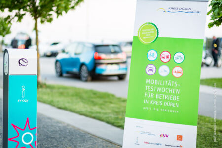 Eine Ladesäule und im Hintergrund ein elektrisch betriebener BMW i3 vor dem Schild der Wirtschaftsförderung im Kreis Düren. E-Mobilität im Unternehmen und Elektromobilitäts-Testwochen im Kreis Düren. Fotograf-Düren-Unternehmensfotografie-Inden-Jülich-Linnich-Langerwehe